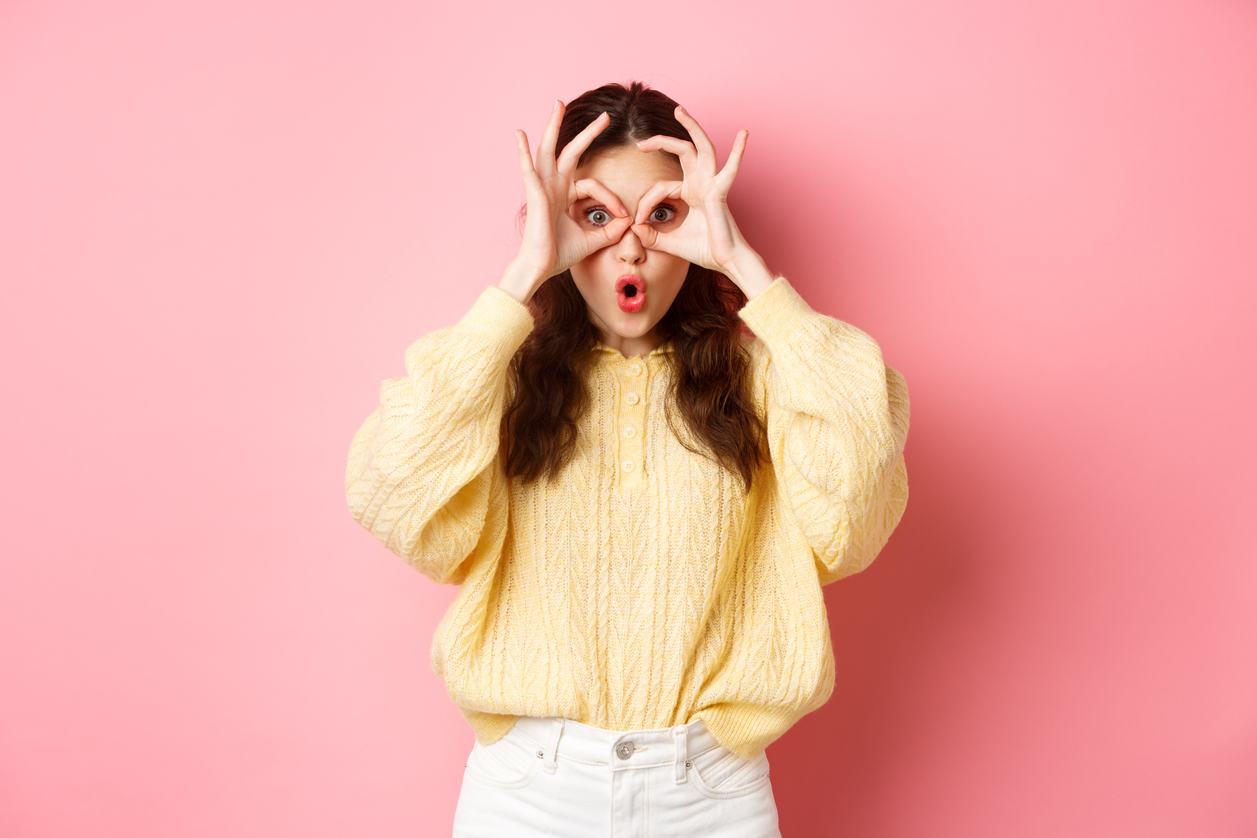 Surprised Young Woman Gasping Amazed, Checking Out Promo Offer, Looking through Hand Binoculars, Standing against Pink Background